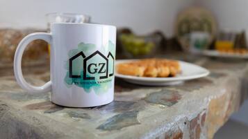 Une tasse blanche ornée d’un logo et d’un texte, posée sur une table en pierre, une assiette avec de la nourriture et un verre en arrière-plan.