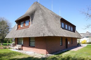 Une maison à toit de chaume, avec une base en briques, un petit porche doté d’un banc et un ciel dégagé.