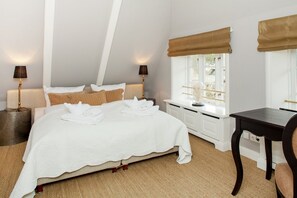 A neatly made bed with white linens and towels, a bedside table with a lamp, a window with a view, and a small desk with a chair.