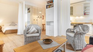 A room with a bed, wicker chairs, a wooden table, a bookshelf, and a kitchen area. - Uaster Reeg III - Studio - Sehr Gepflegte und Geschmackvoll Eingerichtete Ferienwohnungen in (Sylt / OT Archsum)