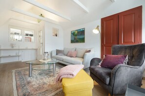 A modern living room with a glass coffee table, a sofa, a yellow ottoman, and a dark armchair. There is a painting on the wall and a red door in the background. - Mootjes Hüs - Gemütliche Obergeschosswohnung Unter Reet mit ca. 50qm Nutzfläche für 2 Personen (Sylt-Ost)