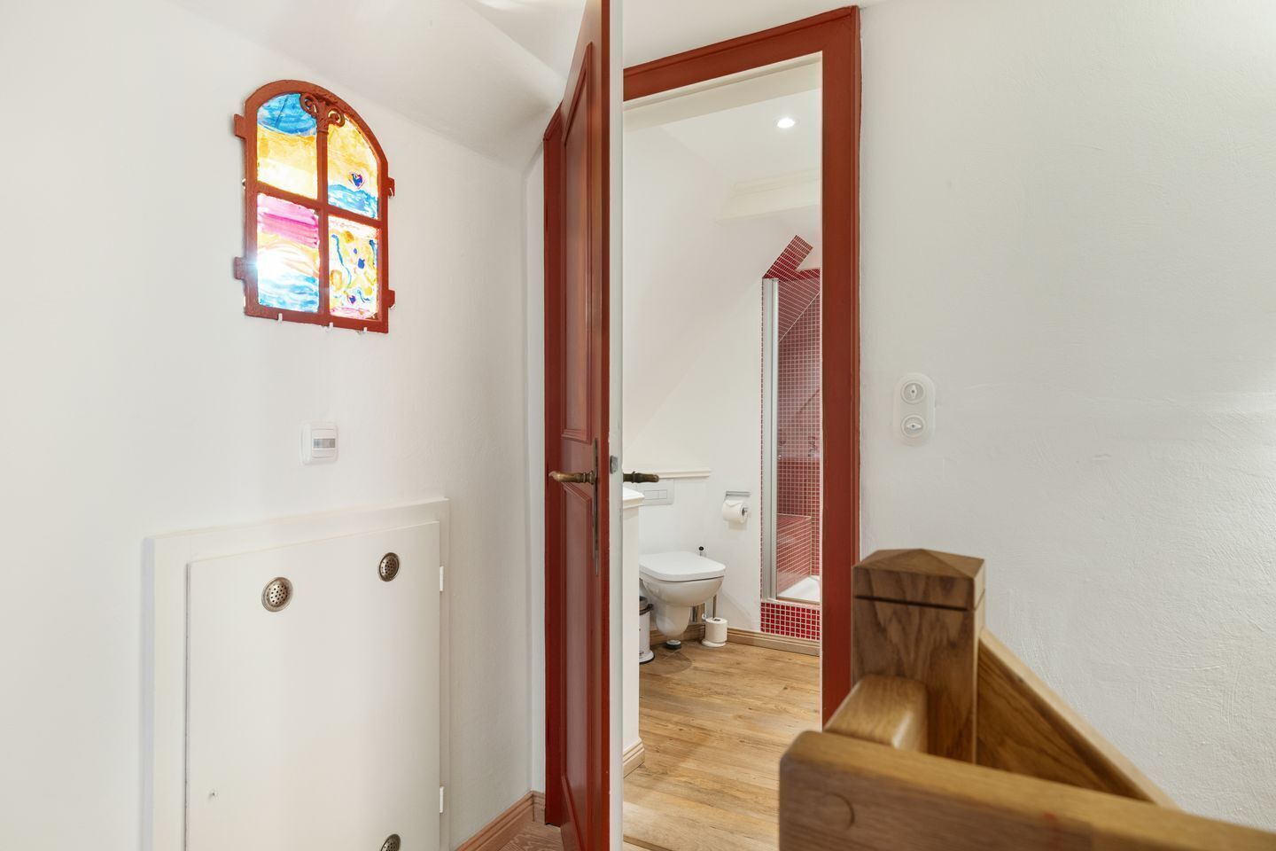 Un couloir avec un escalier en bois, une porte blanche et une fenêtre à vitraux.
