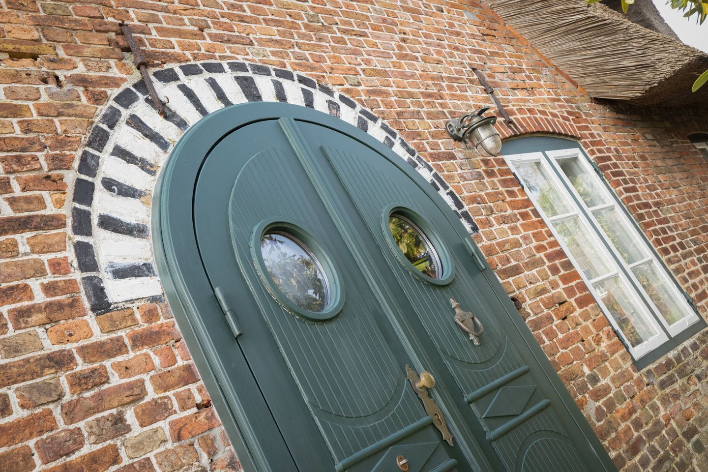 Una porta verde con due finestre circolari e una più piccola sopra, incastonata in un muro di mattoni con un arco decorato a motivi bianchi e neri.