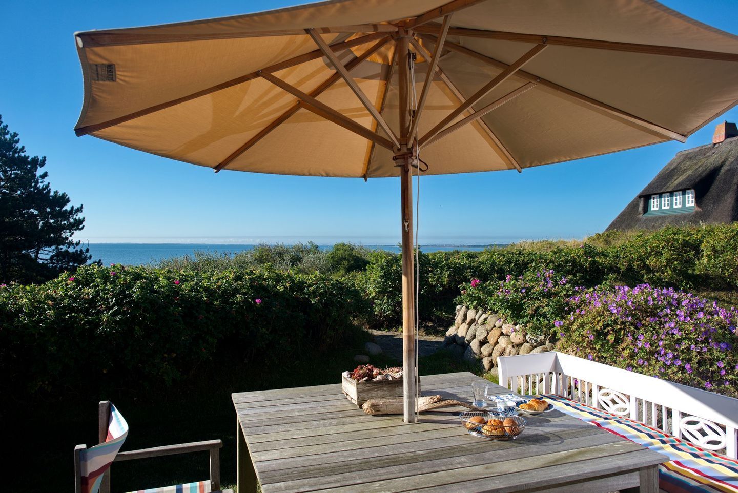 Eine Holzterrasse mit Tisch, Sonnenschirm und Blick auf das Meer sowie ein Haus mit einem Reetdach.