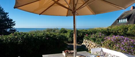 Una terraza de madera con una mesa, una sombrilla y vistas al mar y una casa con techo de paja.