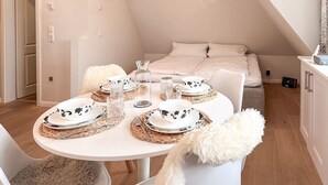 A cozy, well-lit dining area with a round table set for four, white chairs, and a bed in the background.