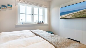 A bedroom with a bed, a window with curtains, a wall-mounted TV displaying a beach scene, and a small shelf with decorative items.
