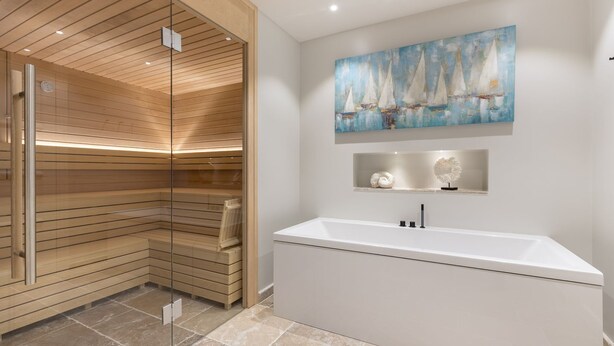 A modern bathroom with a glass-enclosed sauna, a large bathtub, and a nautical-themed painting above the sink.