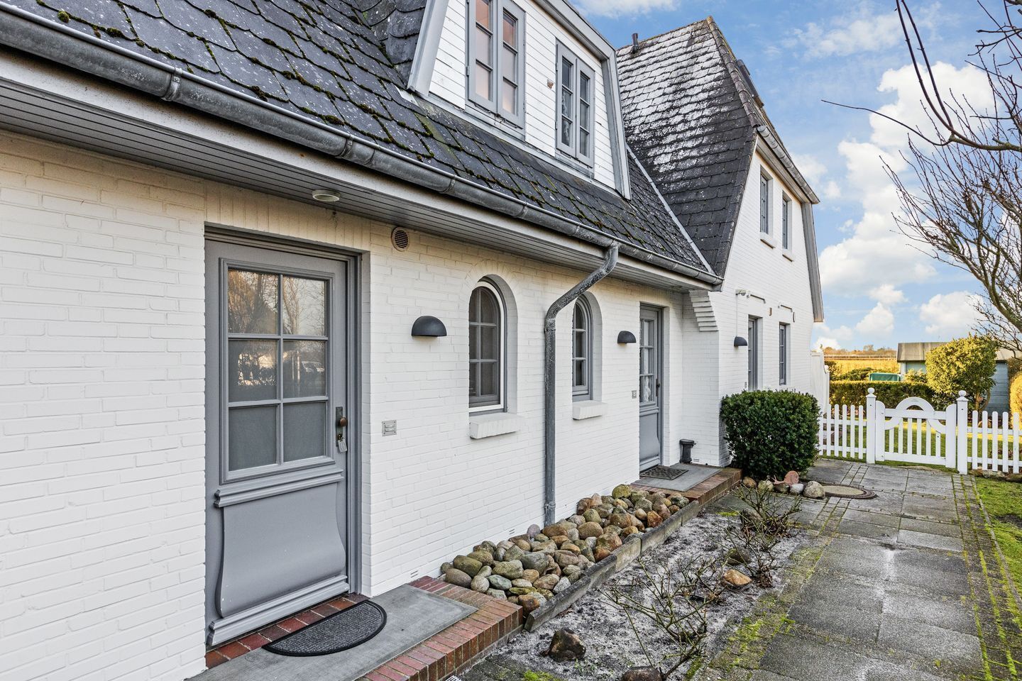 A white brick house with a grey door, a small porch with a stone ledge, and a white picket fence.