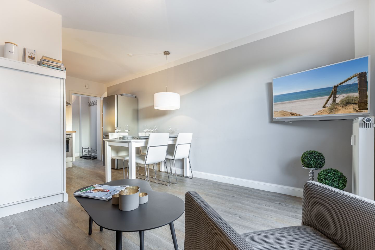 A modern living room with a dining area, a large window with a beach view, and a flat-screen TV mounted on the wall.