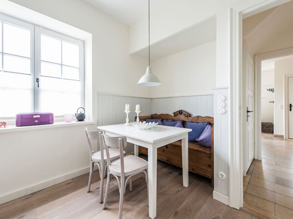 A bright room with a white table and chairs, a wooden bench with cushions, a window with a purple suitcase, and a white pendant light.