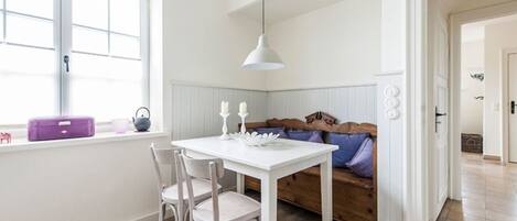 A bright room with a white table and chairs, a wooden bench with cushions, a window with a purple suitcase, and a white pendant light.