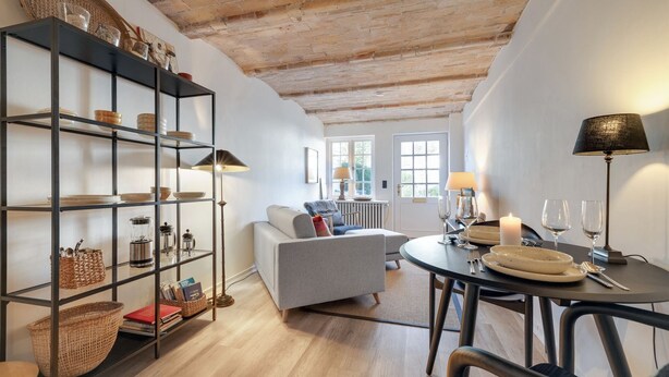 A cozy living room with a fireplace, a dining area with a table set for two, and a bookshelf filled with various items. - Kleine Heimat, Haus Stutenhof Whg. 4 - Die Kleine Heimat Befindet Sich im Herzen von Keitum und (Sylt)