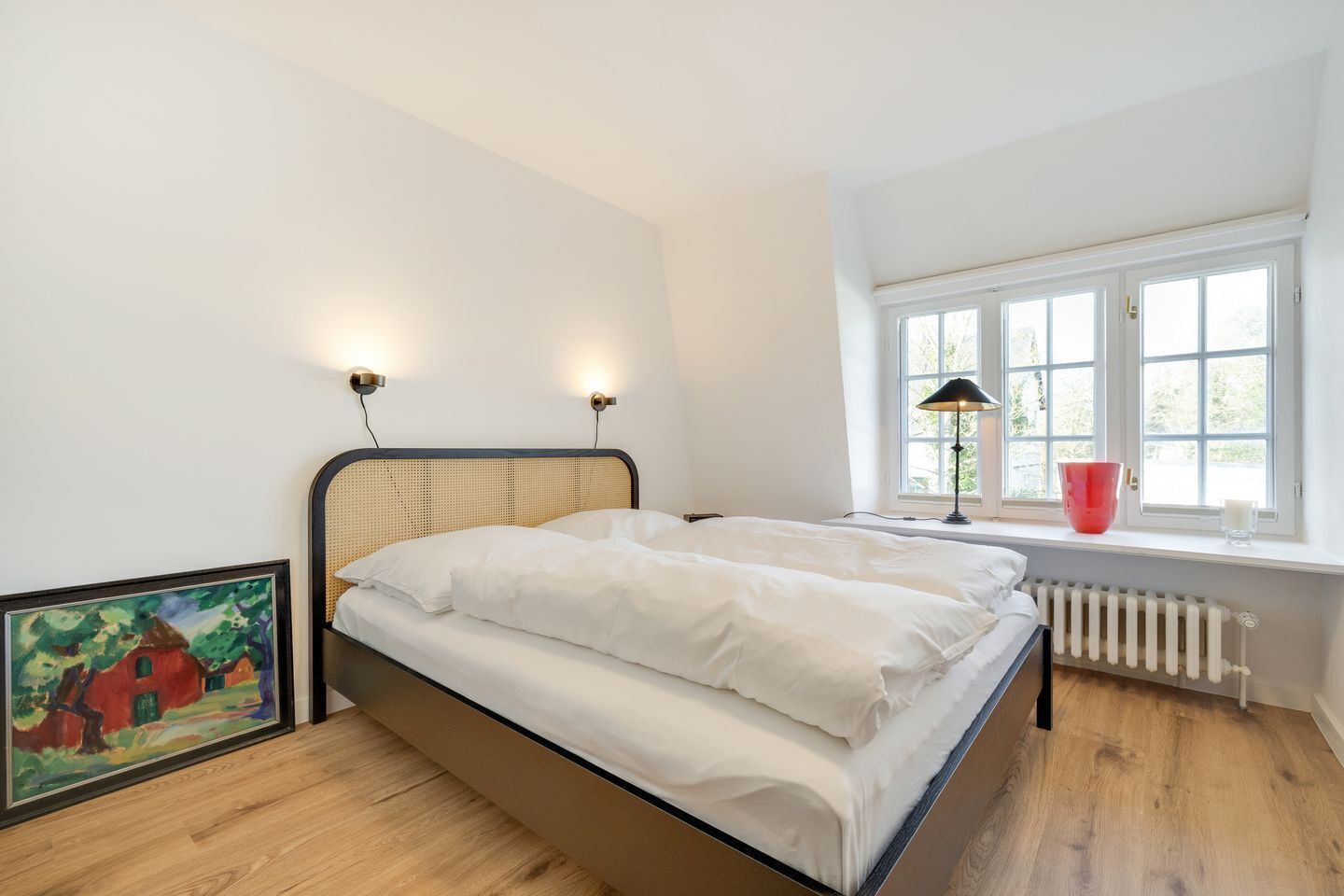 A bedroom with a bed, a radiator, a window with a view, and a painting on the wall.