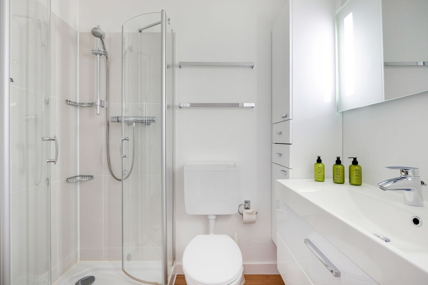 A modern bathroom with a glass shower enclosure, a white toilet, a sink with a mirror above, and three green bottles on the countertop.
