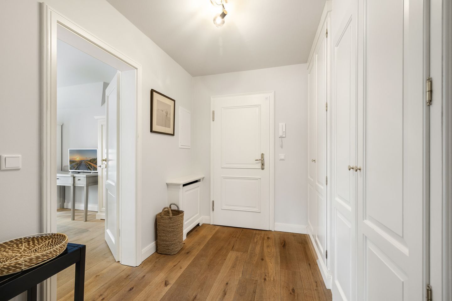 Un couloir aux murs blancs, avec un plancher en bois, un tableau encadré, un panier et une porte.