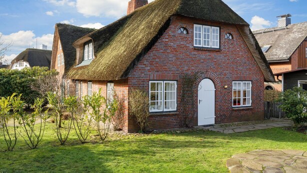 A traditional brick house with a thatched roof, a white door, and a small garden with young trees.