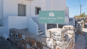 Schild für 'Helen Luxury Villas Naxos' mit einer Steinmauer und einem weißen Gebäude im Hintergrund.