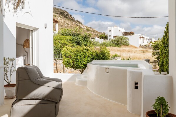 A modern outdoor living area with a sofa, a hot tub, and a view of white buildings and greenery.