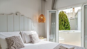 Ett sovrum med en säng, kuddar, ett sängbord, en lampa och utsikt över en balkong med en grön häck.