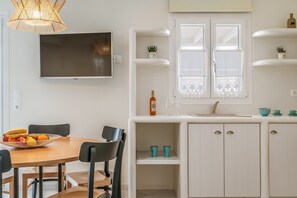 A modern kitchen with white cabinetry, a dining table with chairs, a fruit bowl, a TV mounted on the wall, and a window with decorative lace.