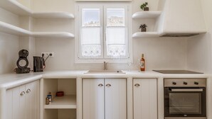 Une cuisine avec des meubles blancs, un four encastré, une fenêtre ornée de rideaux en dentelle et une petite plante posée sur le plan de travail.