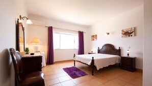 A bedroom with a bed, a chair, a dresser, a lamp, and a window with curtains. - Villa Sol Nascente in Porches (Silves)