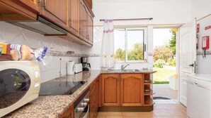 A kitchen with wooden cabinets, a granite countertop, a microwave, a coffee maker, and a view of the garden through a window. - Villa Sol Nascente in Porches (Silves)