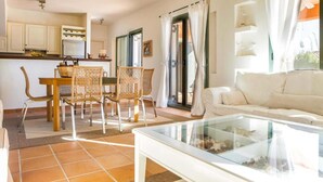 Un salon lumineux avec un canapé blanc, une table basse en verre, des fauteuils en osier, une table à manger et un coin cuisine doté de meubles blancs et d’un réfrigérateur.