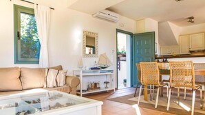 Un salon comprenant un canapé, une table basse avec un plateau en verre, un coin repas avec des chaises en osier, une cuisine avec des placards et une porte donnant sur l’extérieur.