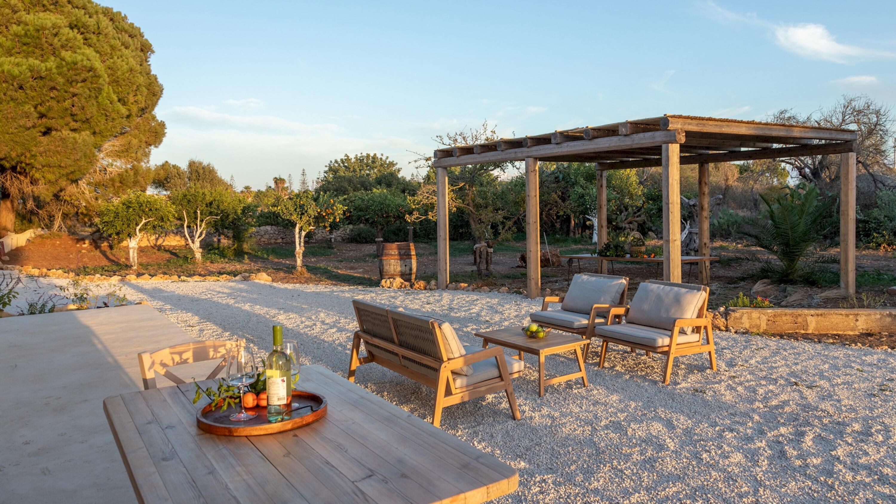 An outdoor seating area with wooden furniture and a pergola, surrounded by gravel and trees.