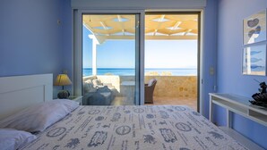 A bedroom with a bed, a nightstand, a lamp, a view of the sea through a glass door, and a beach chair outside.