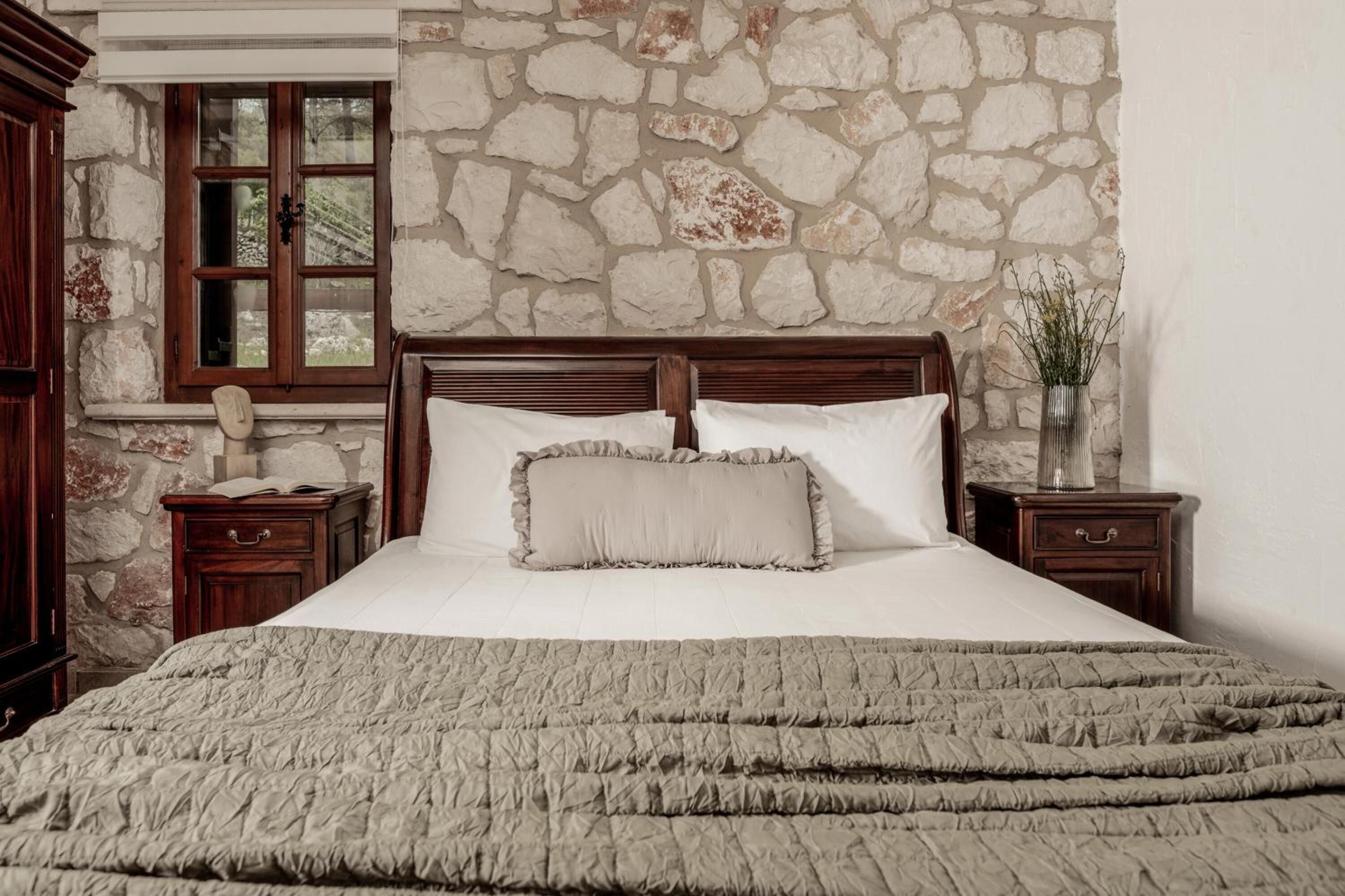 Un dormitorio con pared de piedra, cabecera de madera, cama grande con colcha, dos mesitas de noche y un tocador con un jarrón.