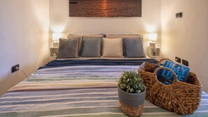 Un lit avec une literie à rayures, deux oreillers, deux lampes de chevet, un tableau accroché au mur représentant un coucher de soleil sur l'eau, et un panier en osier contenant des livres et une plante.