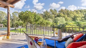 Un espace extérieur aménagé avec des sièges, protégé par un auvent, doté de coussins bleus, d’une table en bois avec un vase et un verre, et offrant une vue sur une piscine et des arbres.