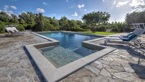 Une piscine rectangulaire aux bords recouverts de carreaux de pierre, entourée de chaises longues et de verdure.