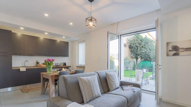 Una cocina moderna con armarios oscuros, una zona de comedor con mesa y sillas, un sofá gris con cojines, y un amplio ventanal con vistas a un jardín.