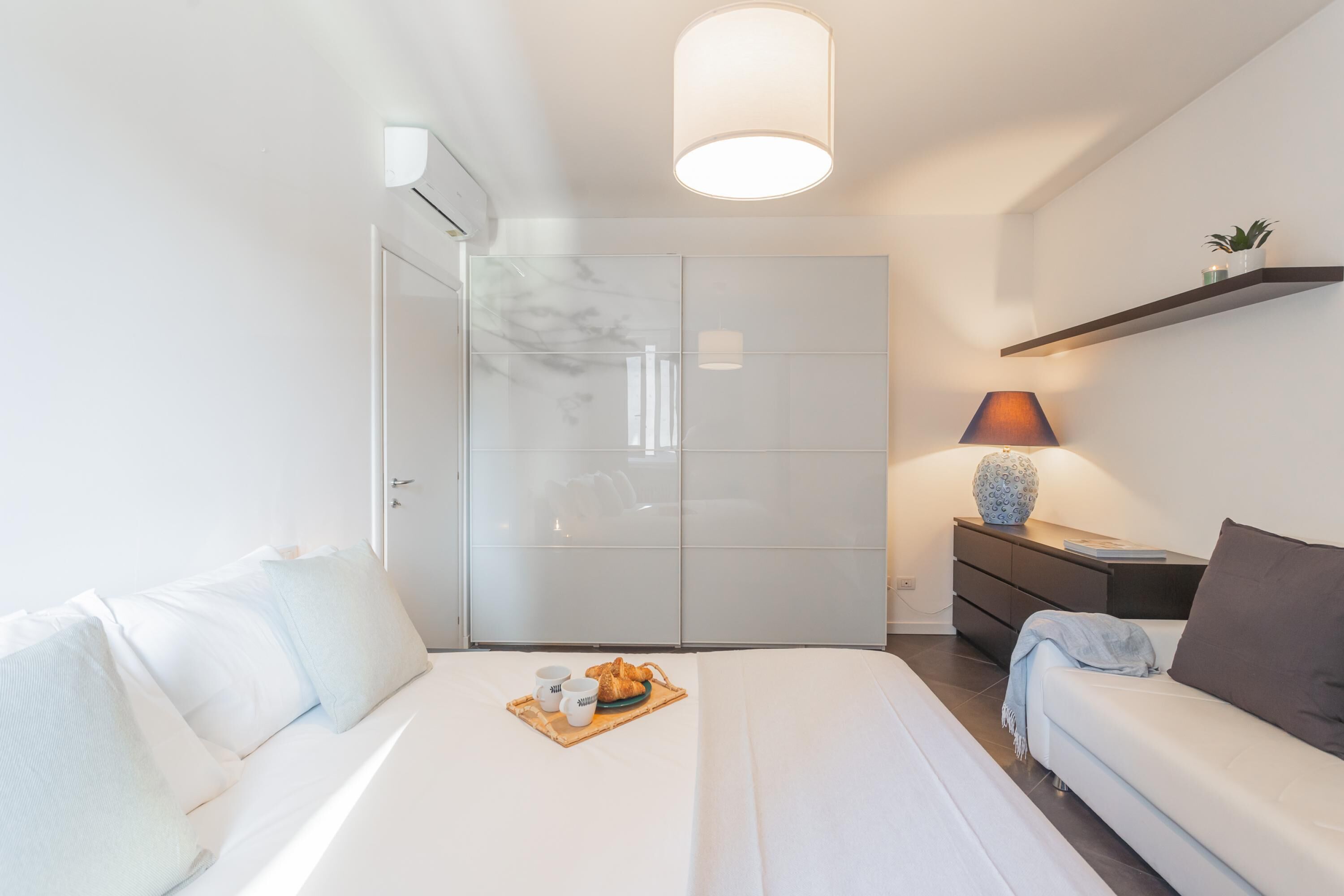 A modern bedroom with a bed, a sofa, a nightstand with a lamp, and a wardrobe.