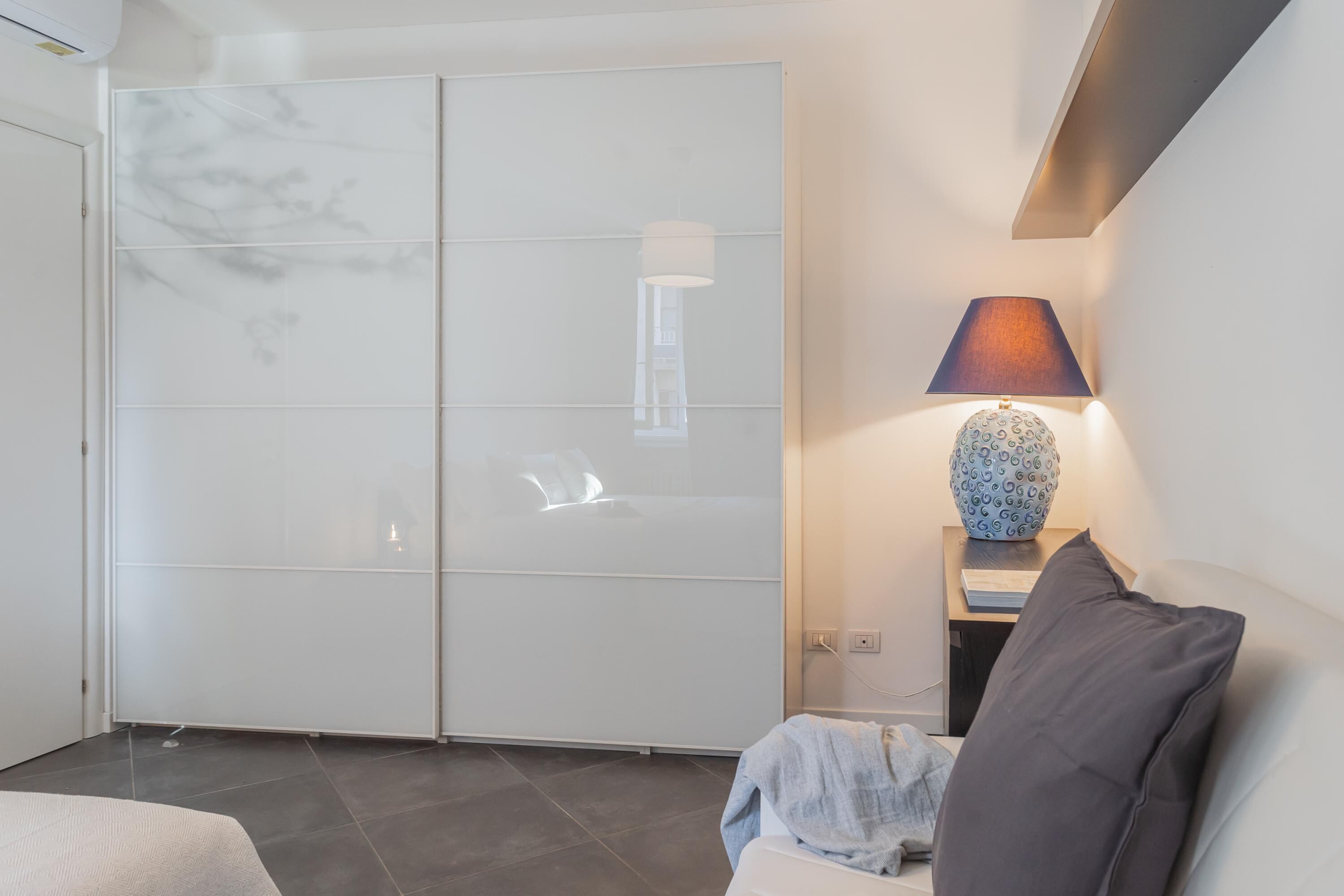 A modern bedroom with a large sliding door closet, a bedside table with a lamp, and a bed with a grey pillow.