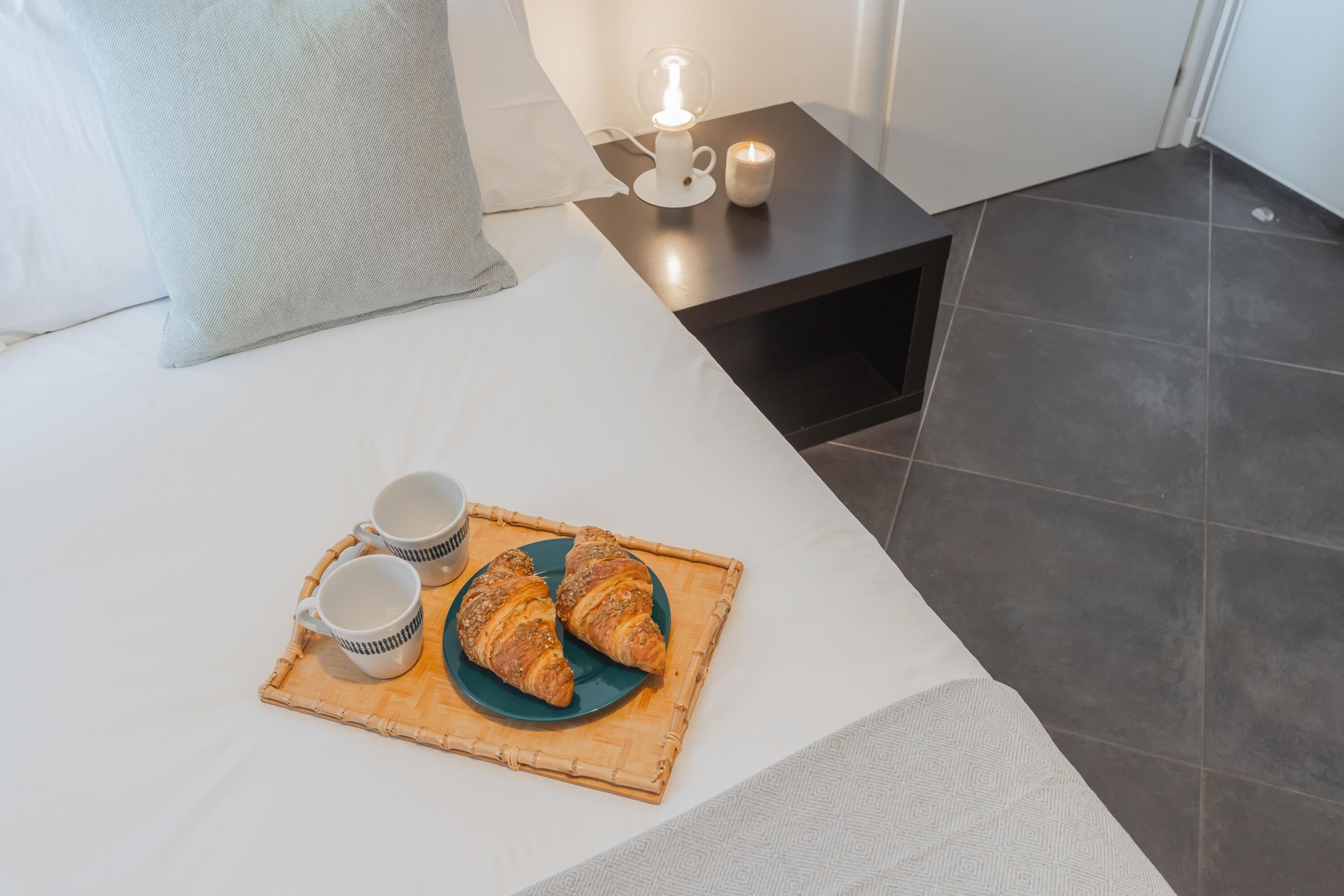 Un lit bien fait, avec un plateau de croissants et deux tasses posées sur un couvre-lit blanc.