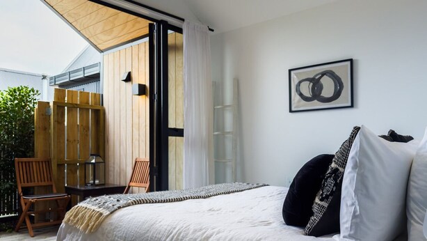 A bedroom with a bed, a wooden chair, a small table, and a view of the outside. - Stunning Matakana Retreat! (Matakana)