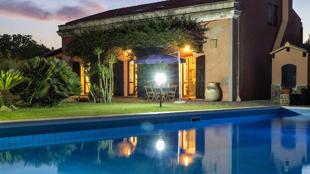 Ein Schwimmbecken mit einer Terrassenanlage, einer überdachten Sitzgelegenheit und einem Gebäude mit einem Ziegeldach.