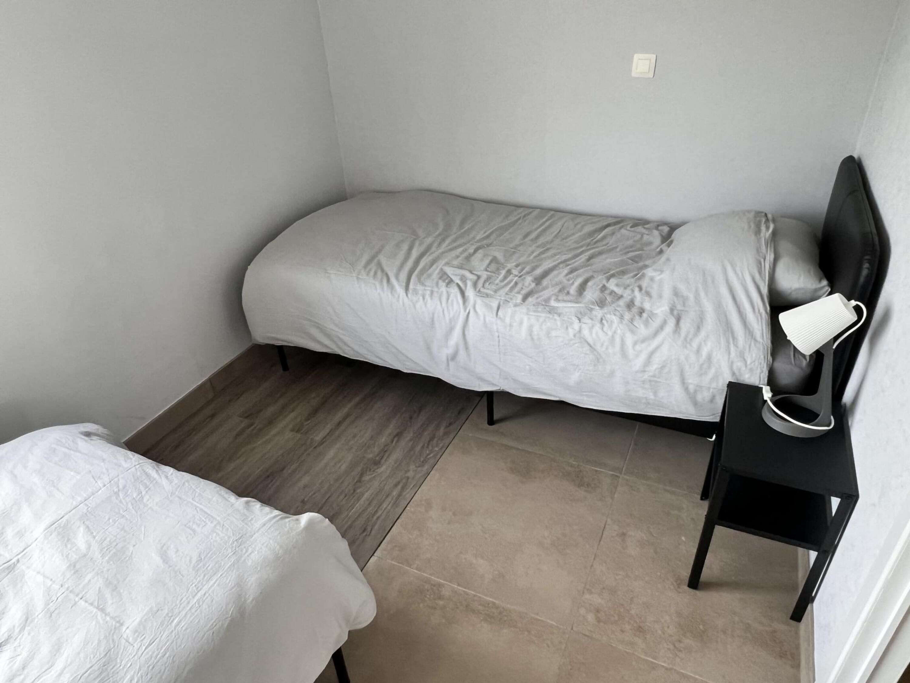 Una camera piccola con un letto singolo, pavimento in legno, un comodino con lampada e una presa elettrica.