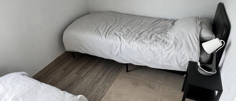 A small, single-bed room with a wooden floor, a bedside table with a lamp, and a wall socket.