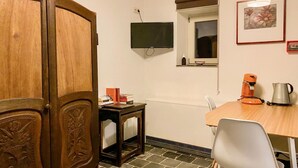 A small, cozy room with a wooden wardrobe, a small table with a chair, a television mounted on the wall, and a window with a view of the outside.