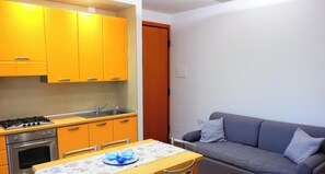 Une cuisine de taille réduite avec des placards jaunes, une cuisinière à gaz et un évier. On y trouve une table à manger avec un saladier bleu. Un canapé gris est placé contre le mur.
