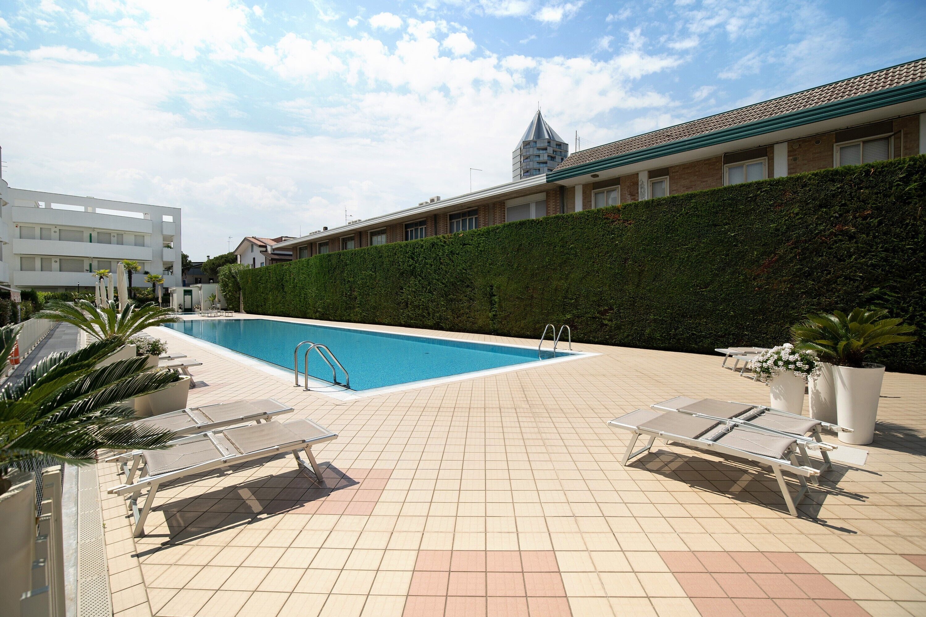 Una piscina con sillones reclinables, una terraza con baldosas y un edificio con un seto verde al fondo.