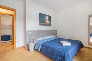 Une chambre à coucher comprenant un lit, une table de chevet, une poubelle, un tableau représentant une scène de plage et une armoire.