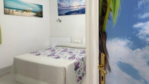 A bedroom with a bed, two wall-mounted paintings, a door with a palm tree mural, and a white ceiling.