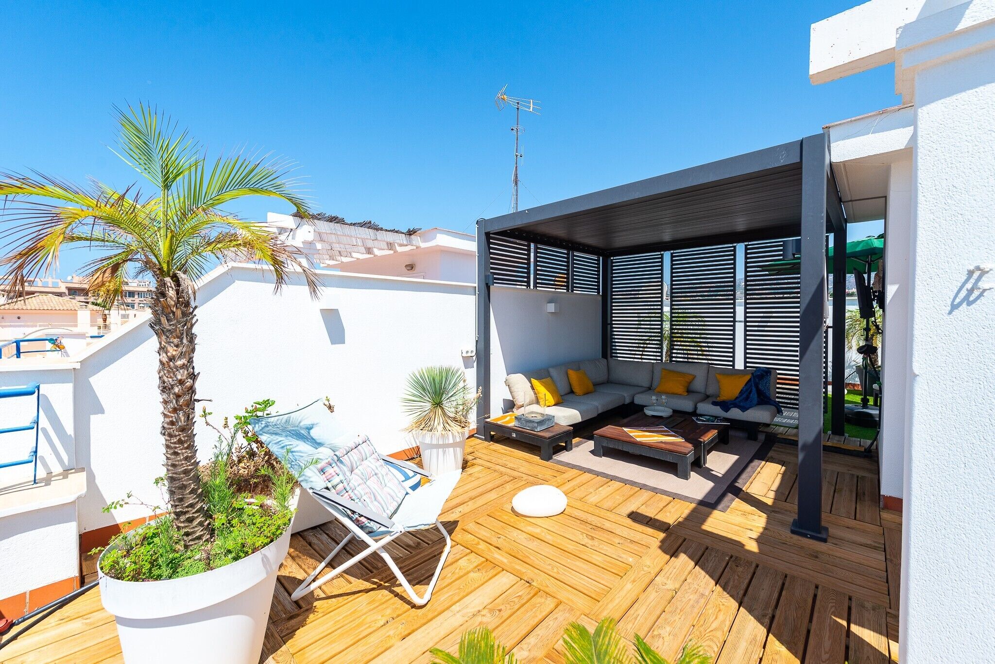 Une terrasse sur le toit avec une plateforme en bois, un coin salon avec un canapé et des coussins, un palmier et une plante en pot.